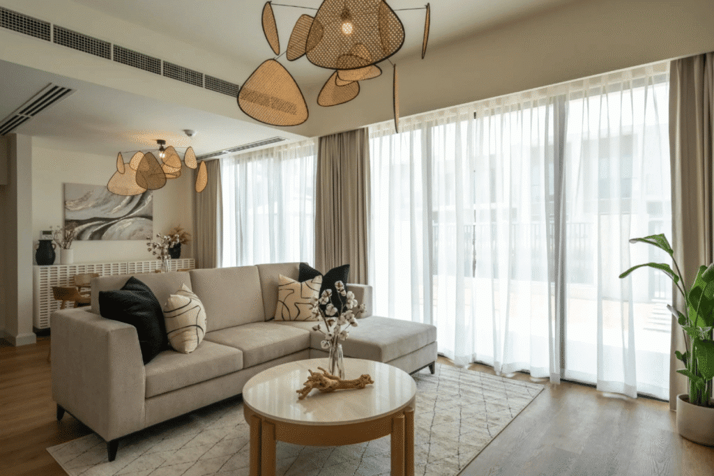 Modern living room with beige sofa, abstract lighting, and large windows with sheer curtains. Minimalist decor and plants.