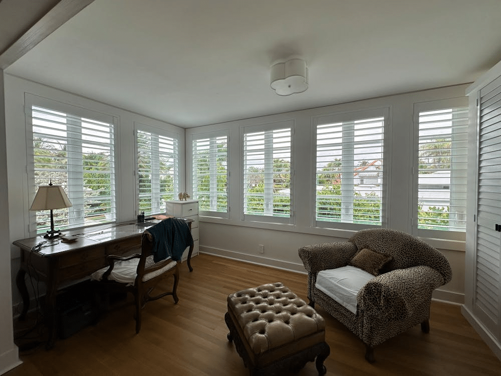 Bright home office with wooden desk, plush chair, and large windows with white shutters offering garden views.