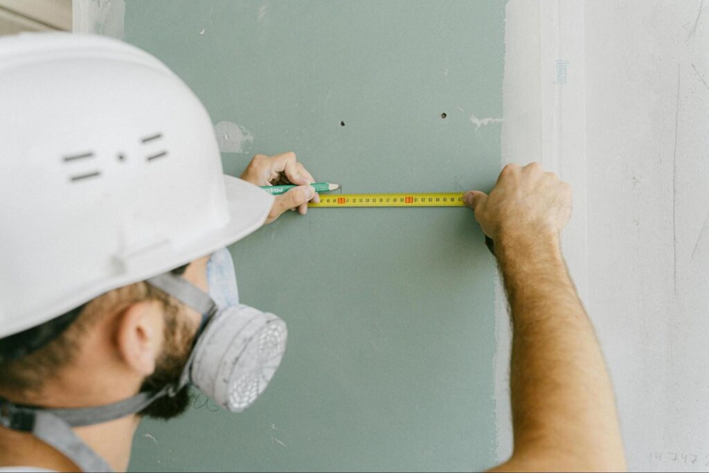 image1 Construction worker measures wall with tape measure while wearing a hard hat and mask. | Sky Rye Design Construction worker measures wall with tape measure while wearing a hard hat and mask.