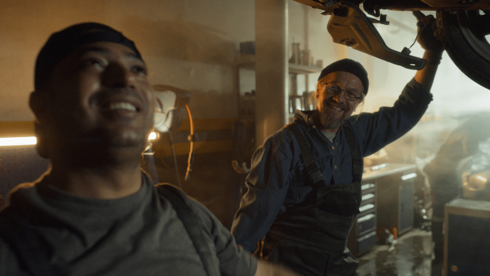 Mechanics smiling while working in an auto repair shop, inspecting the undercarriage of a car.