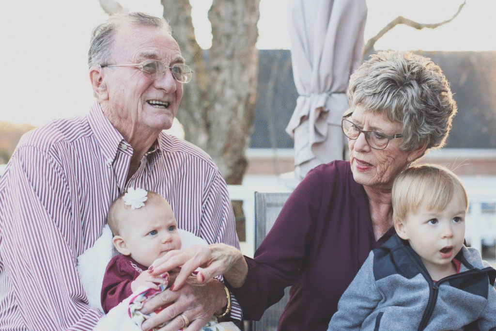 Elderly couple smiling with grandchildren, a baby and a toddler, enjoying family time outdoors.