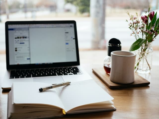 Open laptop and notebook with pen on a wooden desk, beside a coffee mug and small flower vase.