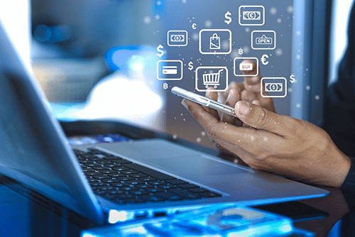 Person using smartphone for online shopping beside laptop, surrounded by digital shopping icons and currency symbols.
