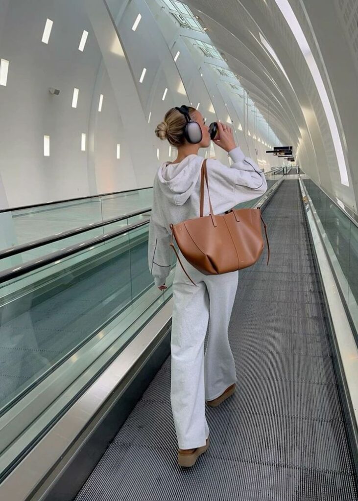Person in gray outfit on airport travelator, wearing headphones and holding a brown tote bag, modern architecture.