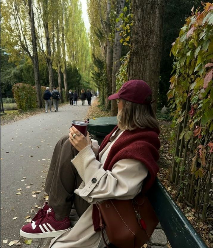 Woman in autumn outfit sitting on park bench with coffee, wearing maroon cap and scarf, surrounded by trees and leaves. Autumn outfit