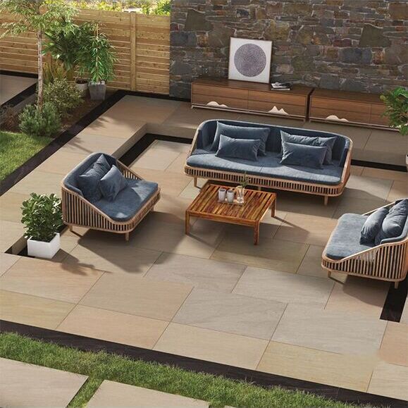 Modern patio with wicker furniture, blue cushions, and a wooden coffee table, surrounded by plants and a stone wall.