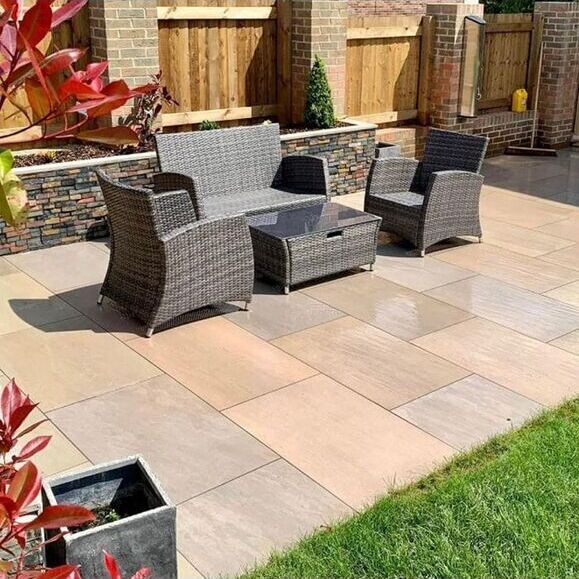 Cozy outdoor patio with wicker furniture set on stone tiles, surrounded by wooden fencing and lush greenery.
