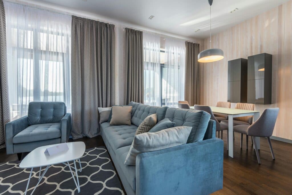 Modern living room with blue sofa, armchair, and dining area. Bright space with large windows and stylish decor.
