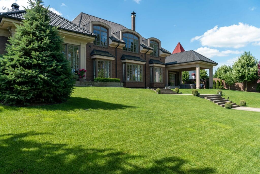 Large luxury brick house with manicured lawn, tall evergreen tree, and clear blue sky.