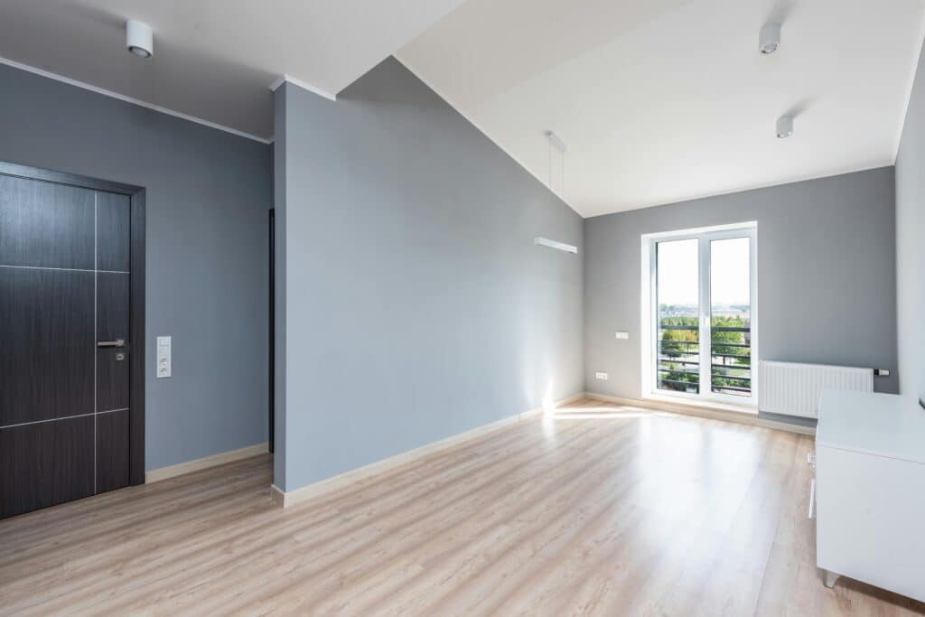 Modern empty room with gray walls, light wood flooring, large window, and natural light. Ideal for living space or office.