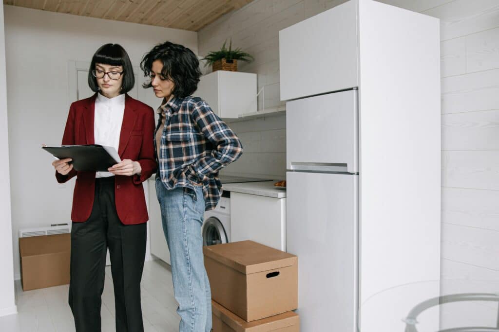 Two women reviewing documents in a modern kitchen with moving boxes, indicating a house move or real estate transaction.