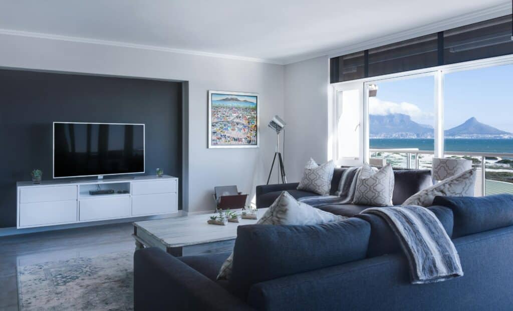 Modern living room with blue sofa, TV, and sea view through large window, featuring elegant decor and cozy cushions.