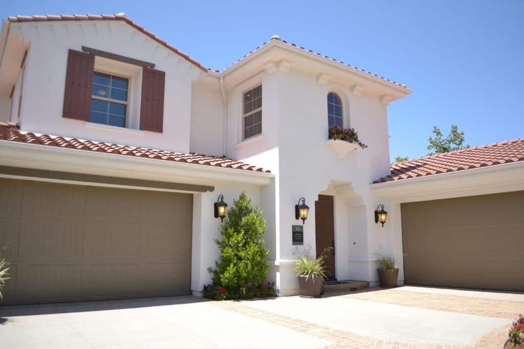 Modern stucco house with red tile roof, twin garages, and landscaped front yard under clear blue sky.