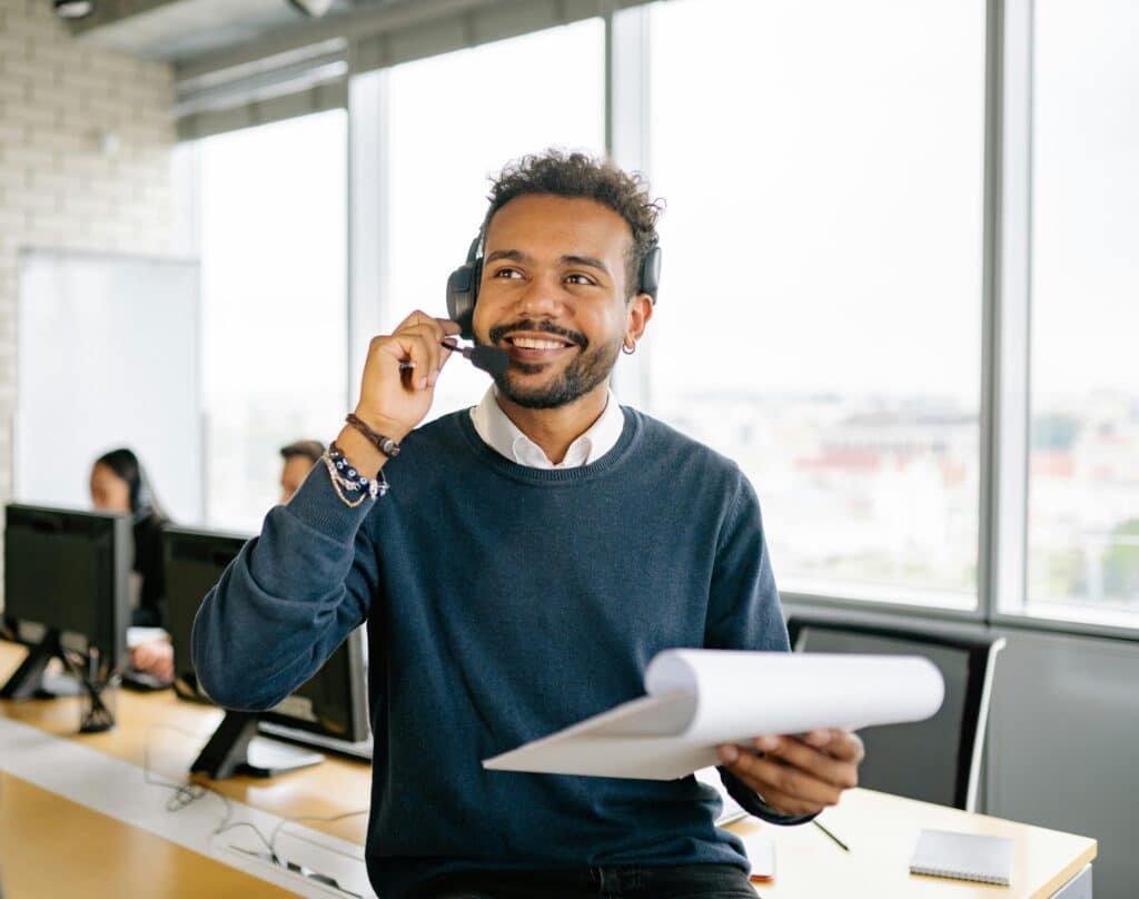 Smiling call center agent with headset, holding a notepad, in a modern office environment.