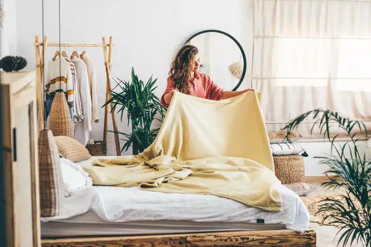 Woman tidying up bed in cozy, plant-filled bedroom with mirror and natural light.