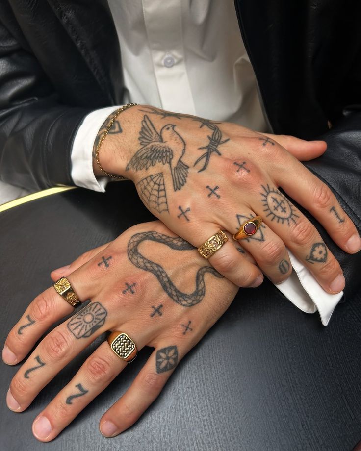Tattooed hands with gold rings and intricate designs, including a bird and snake, resting on a table.