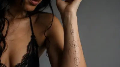 Close-up of person's forearm with minimalist geometric black-ink tattoo of stacked circles and lines, lace bralette visible