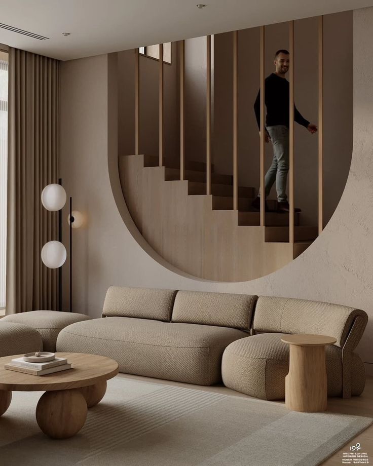 Modern living room with beige furniture, round coffee table, vertical staircase, and man descending stairs through a circular opening.