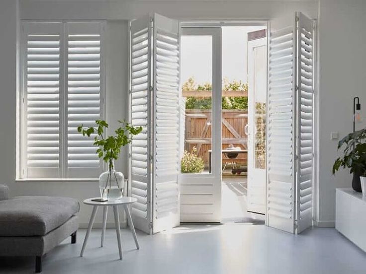 Bright room with open white shutters and door leading to a sunny patio, featuring modern decor and indoor plants.