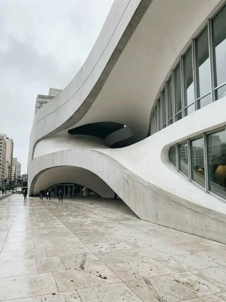 Curving modern concrete cultural building with large windows and open plaza on rainy day, pedestrians