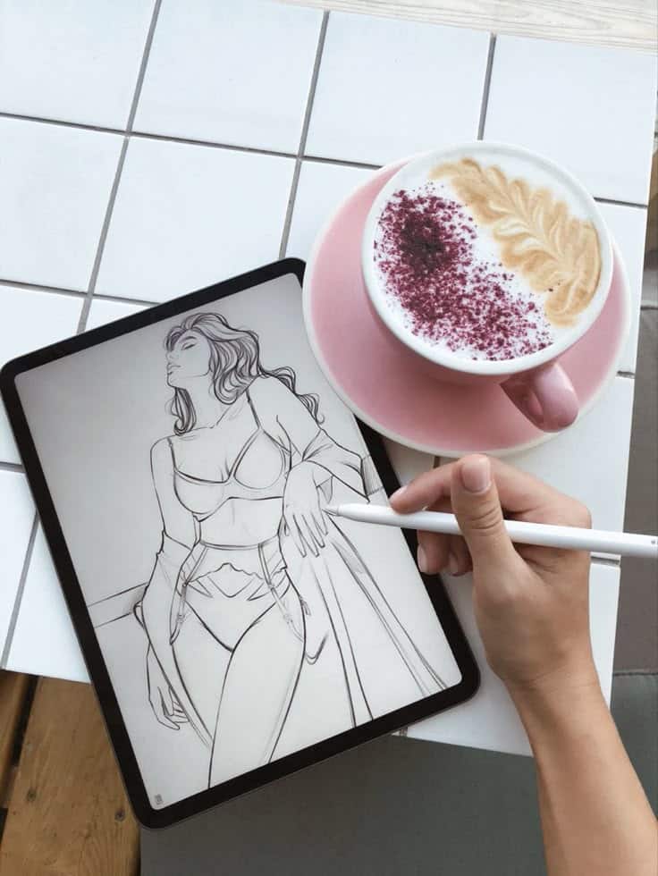 Digital drawing on a tablet next to a cappuccino with latte art in a pink cup.
