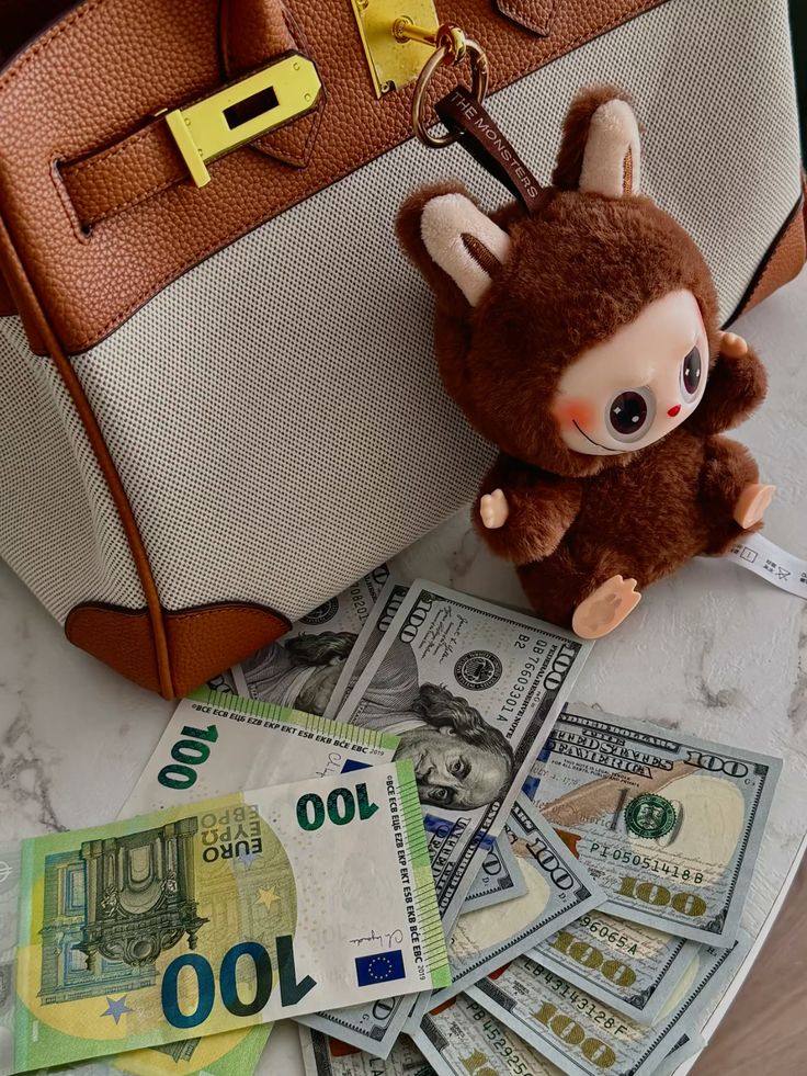 Luxury handbag with plush toy, euro, and dollar bills on marble surface.