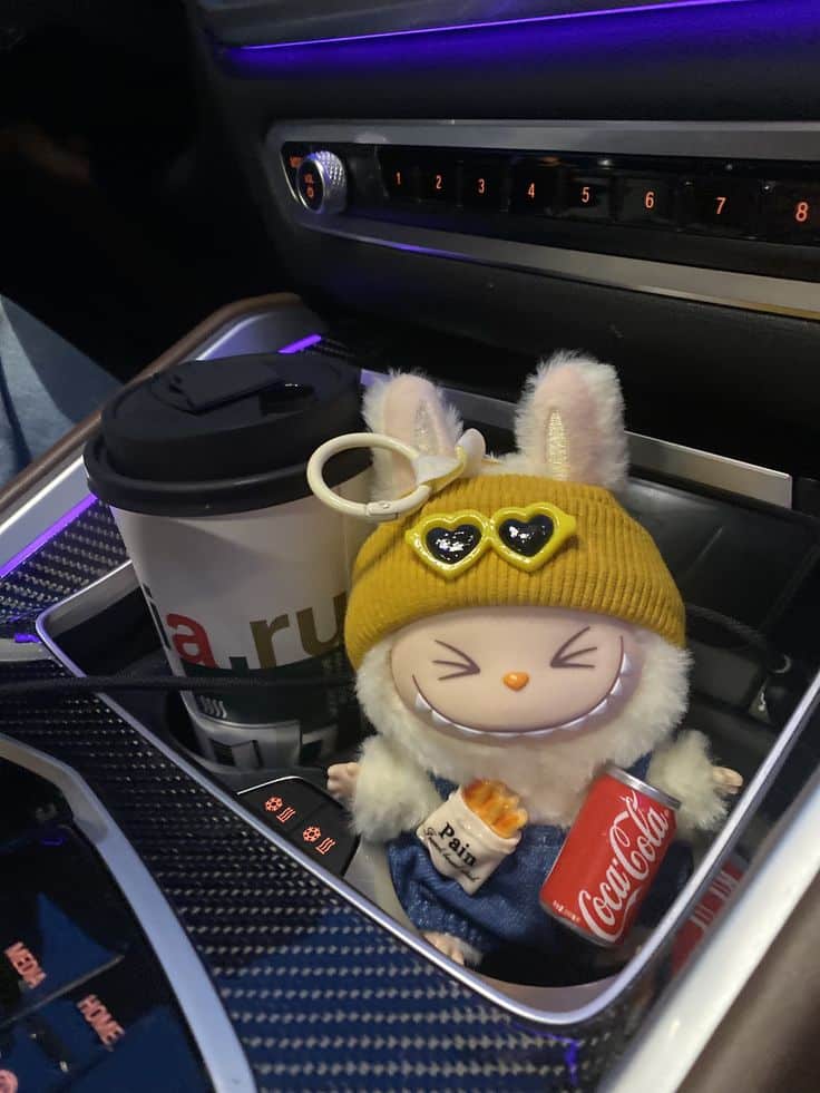 Cute toy bunny with yellow hat and coffee in a car cup holder, holding a mini Coke can, in a dimly lit car interior.