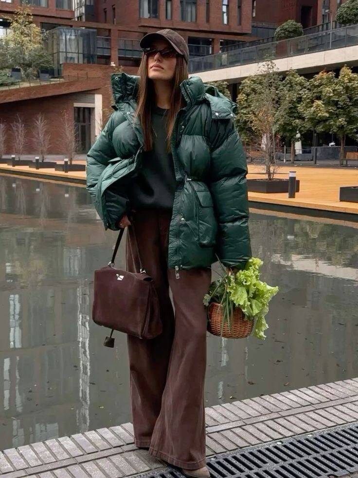 Woman in green puffer jacket and hat, holding a basket of greens and a brown bag by urban canal.