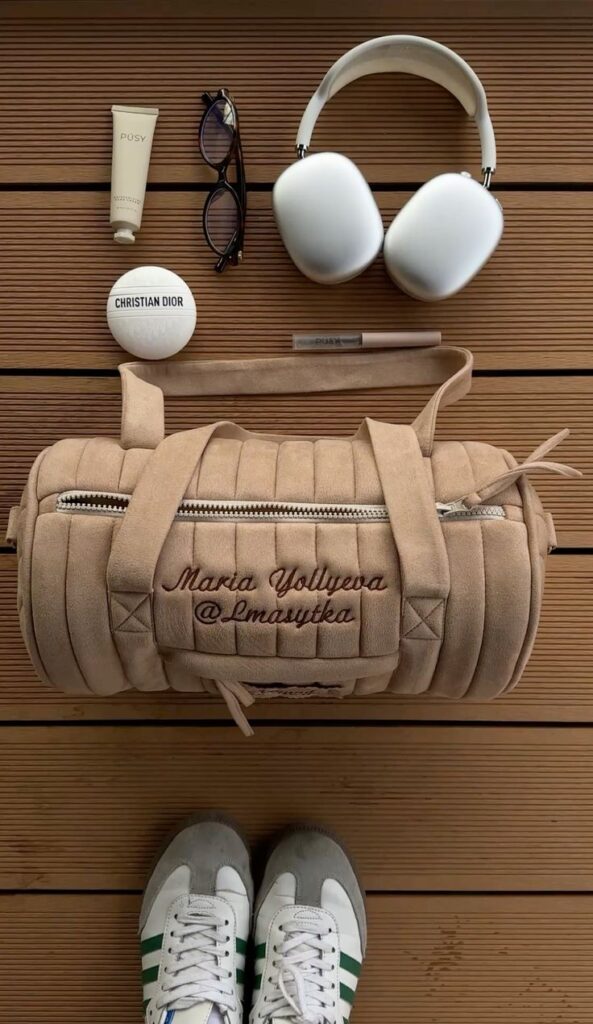 Travel essentials on wooden surface: suede duffle bag, headphones, glasses, sneakers, beauty products.