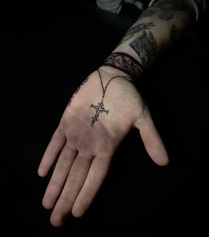 Close-up of a hand with a detailed cross tattoo on the palm, surrounded by other intricate tattoos, against a dark background.
