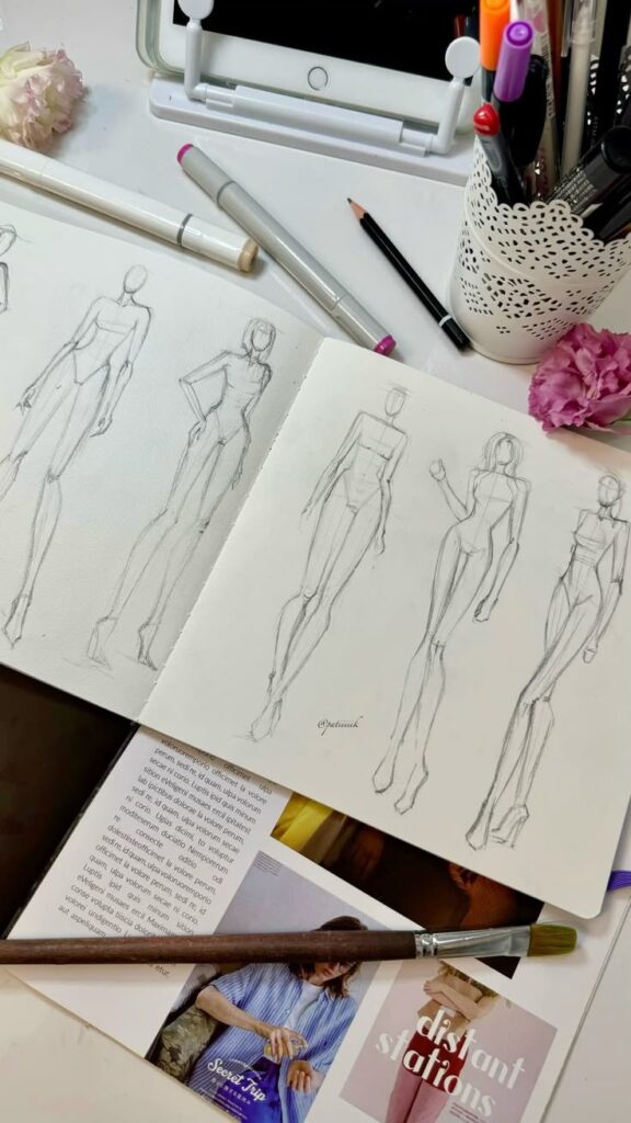 Fashion sketchbook with figure drawings on a desk surrounded by art supplies and flowers.