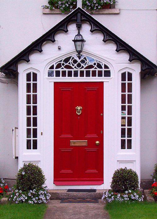 Charming red front door with decorative glass panels, surrounded by flowers and greenery.