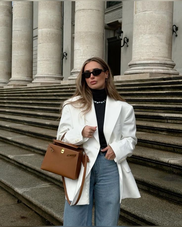 Fashionable woman in white blazer, sunglasses, and brown handbag on stone steps. Elegant urban style.