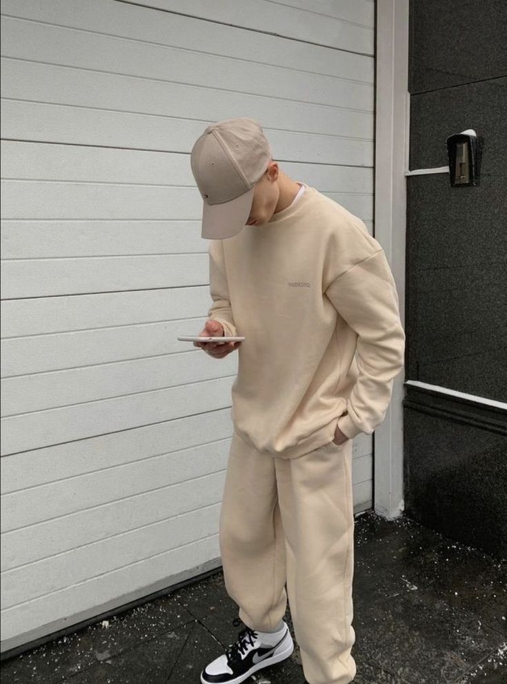 🐼 Casual beige outfit with matching cap and sneakers, person checking phone against a white garage door background. | Sky Rye Design Casual beige outfit with matching cap and sneakers, person checking phone against a white garage door background.