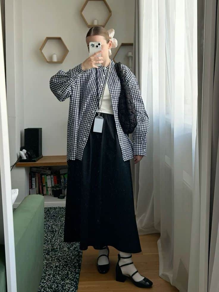Person in checkered shirt and black skirt takes a mirror selfie, showcasing a casual, stylish outfit.