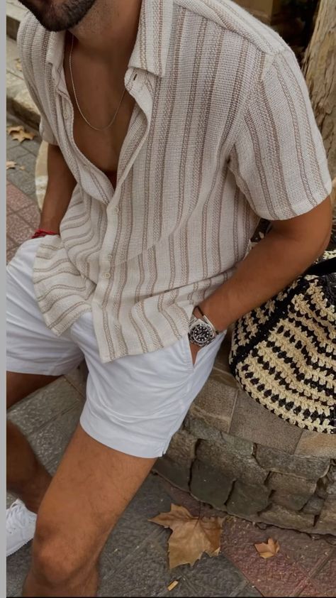 Stylish man in striped shirt and white shorts sitting on a stone bench, carrying a woven bag, showcasing summer fashion.