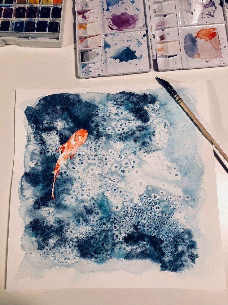 Watercolor painting of an orange koi fish swimming in blue water with paint palettes and brush nearby.