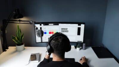 Person working on desktop setup with dual monitors, headphones, desk lamp, and potted plants in a modern workspace.