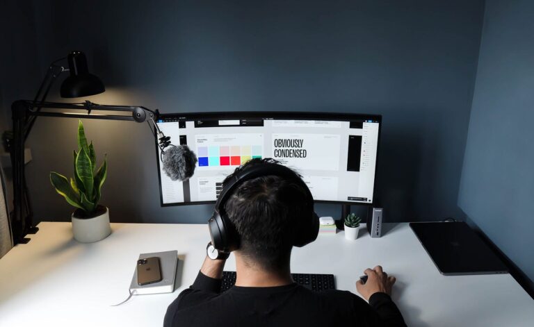 Person working on desktop setup with dual monitors, headphones, desk lamp, and potted plants in a modern workspace.