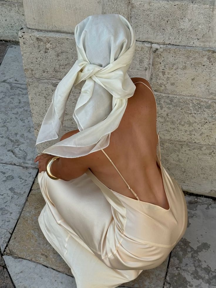Woman in elegant backless dress and headscarf crouched by stone wall, showcasing chic summer style.