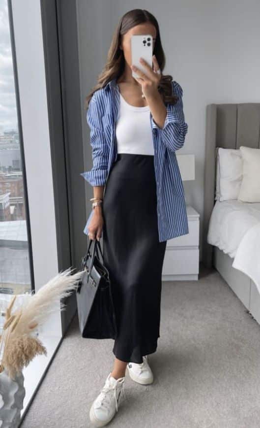 Woman in a striped shirt and black skirt taking a mirror selfie in a modern bedroom with large windows.