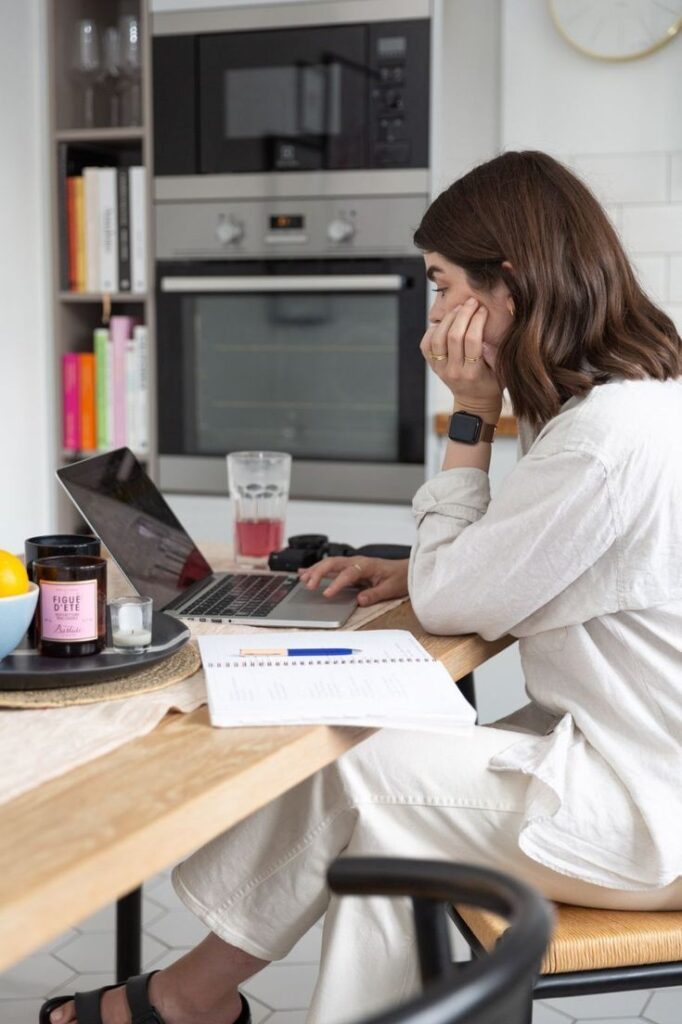 Woman working on a laptop at a kitchen table, surrounded by a notebook and candle.