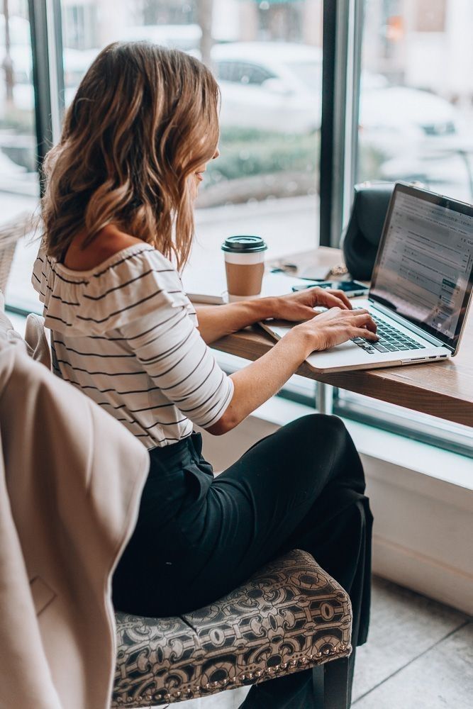 Woman working on a laptop in a cafe with coffee next to her, wearing a striped shirt and black pants, near a large window.