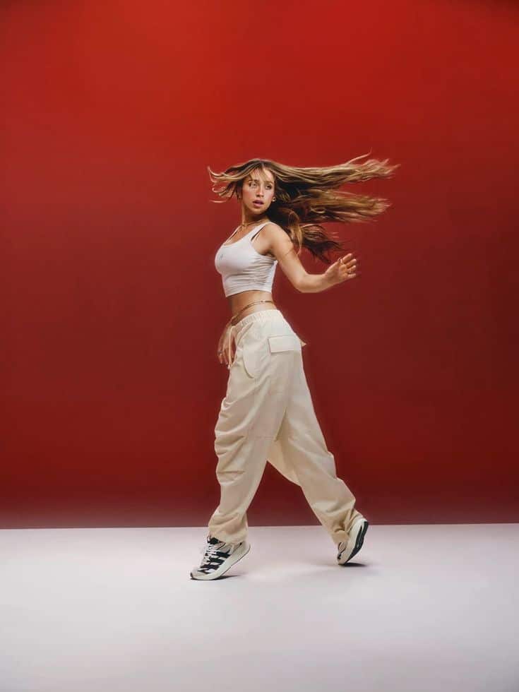 Dancer in motion with flowing hair, wearing a white crop top and pants, poses against a vibrant red background.