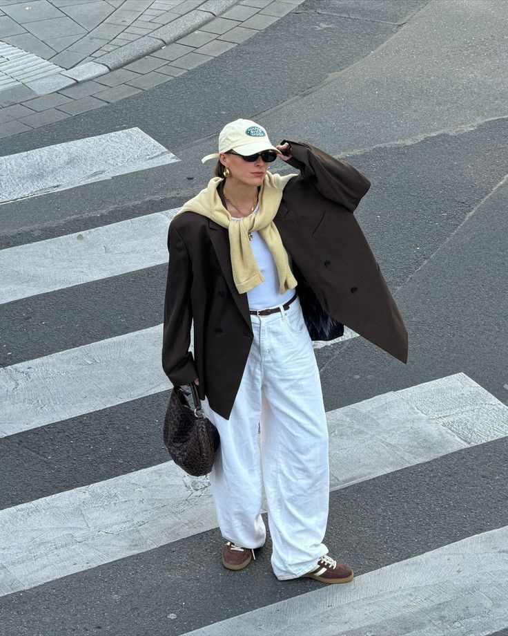 Fashionable person in oversized blazer, white pants, and sneakers crossing street at crosswalk, wearing cap and sunglasses.