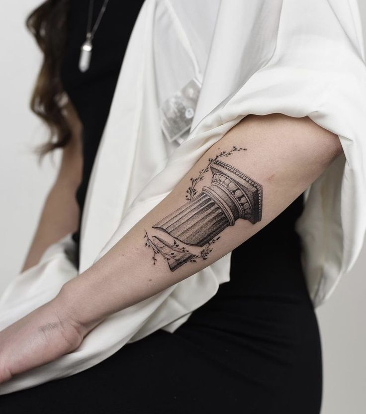 Tattoo of a classical column with floral accents on a person's forearm, draped in white fabric.