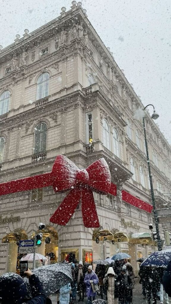_ 1 Snowy city street with a grand building decorated with a large red bow, bustling with people under umbrellas. | Sky Rye Design Snowy city street with a grand building decorated with a large red bow, bustling with people under umbrellas.