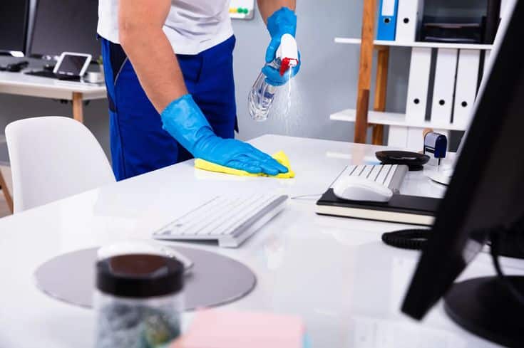 Person cleaning office desk with spray and cloth for hygiene and organization.
