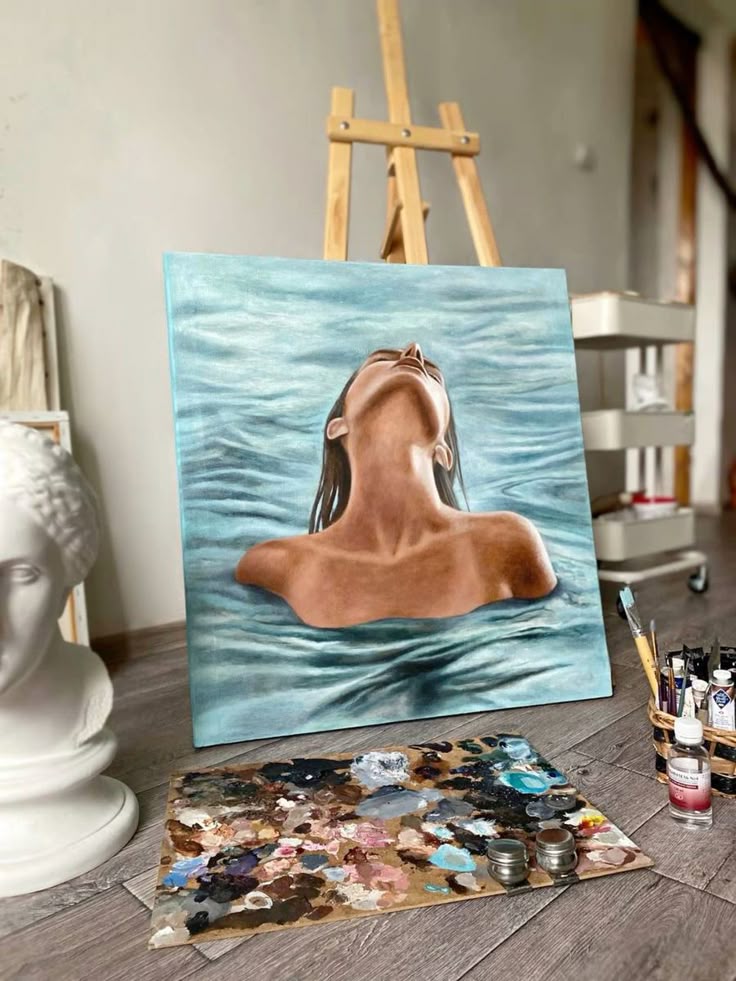 Painting of a person in water on an easel, surrounded by art supplies and a sculpture bust in an artist's studio.