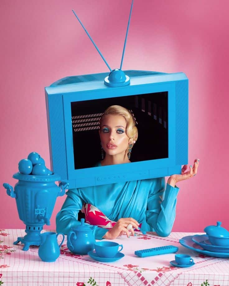 Vintage TV head woman in blue, sitting at a colorful table with retro style decor against a pink backdrop.